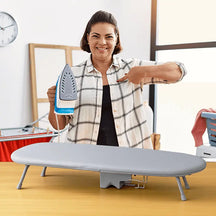Woman holding an iron next to a portable and foldable ironing board in a small home setting.